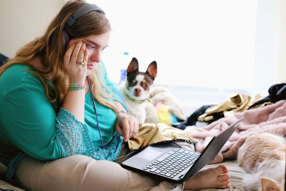 A woman wearing headphones looking at a laptop with a dog beside her